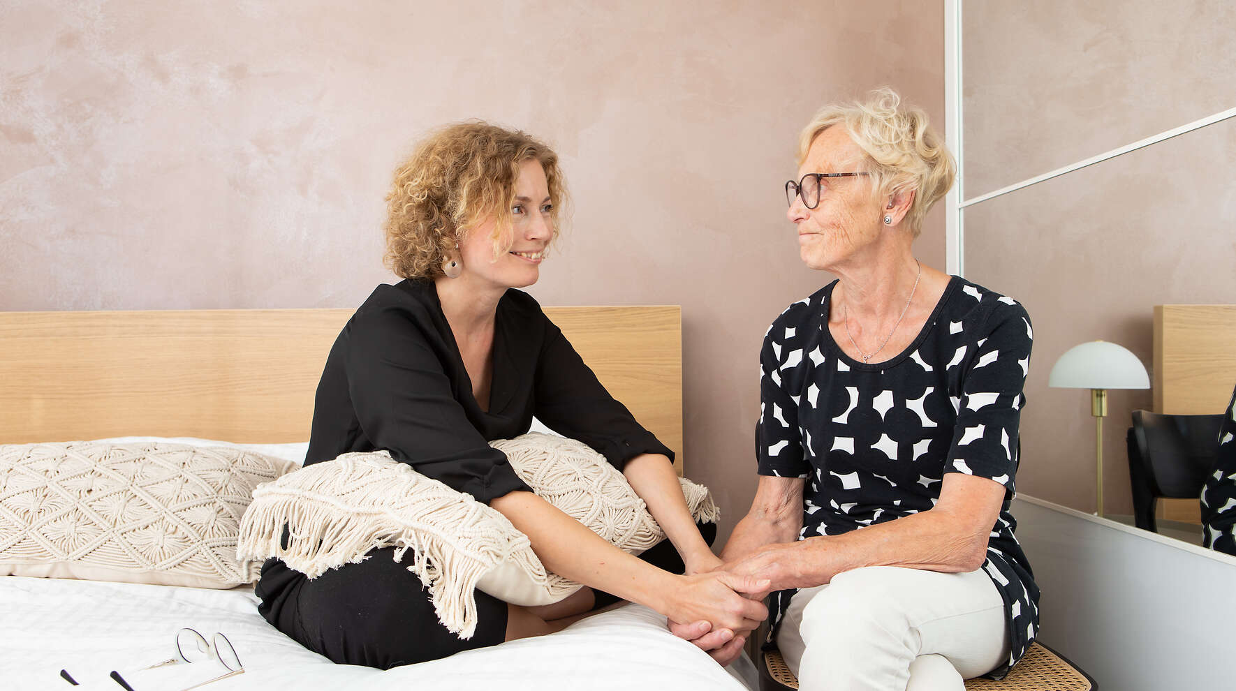 Tytär ja hänen iäkäs äitinsä istuvat vierekkäin. He katsovat toisiaan hymyillen käden yhteen liitettyinä.