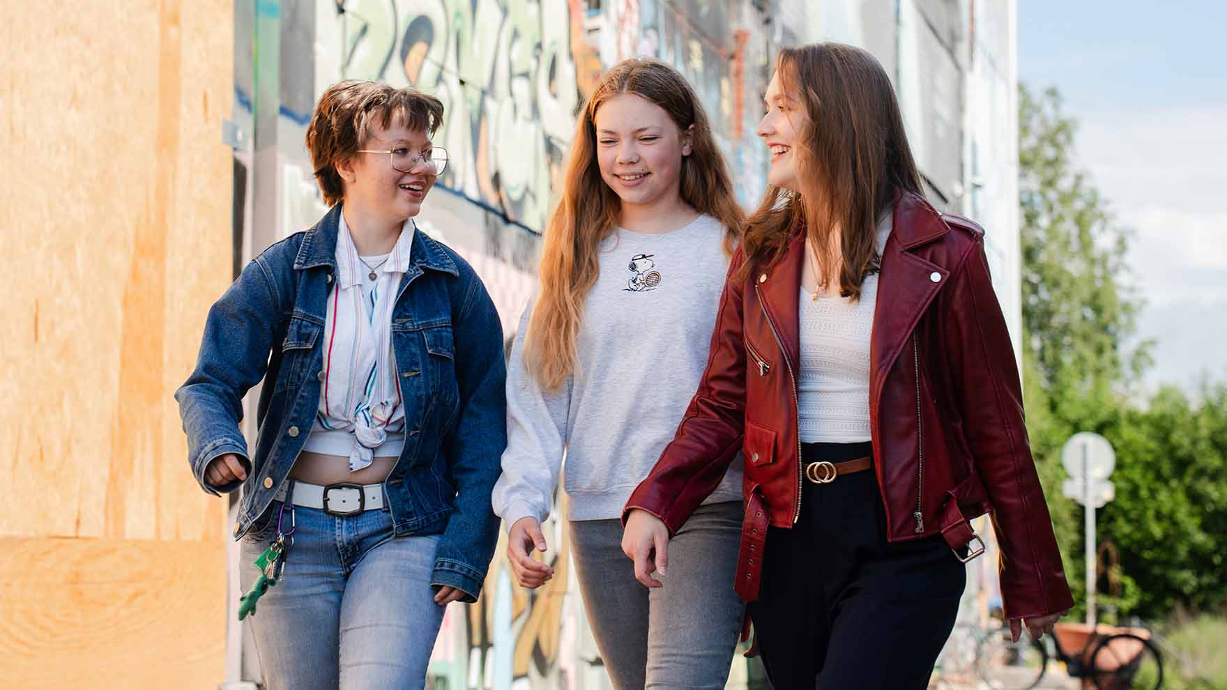 Three young women are walking side by side. In the background, you can see a wall covered in graffiti and some trees.