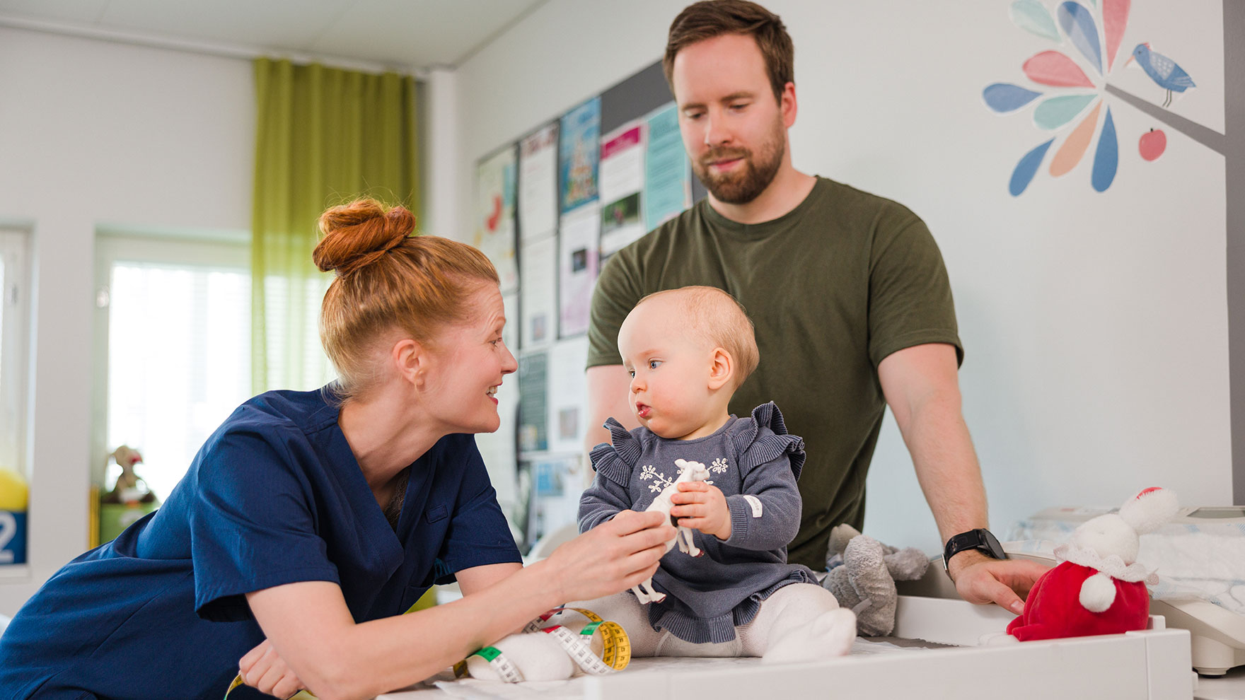 A nurse at a child care clinic is talking to a baby who is sitting on a table. The father is standing behind the baby.