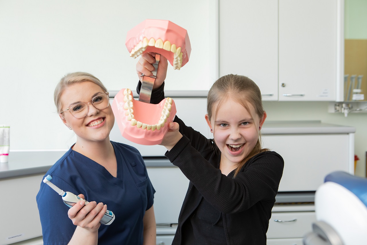 Tyttö pitelee käsissään hampaistomuottia ja hänen vieressä suun terveydenhuollon ammattilainen hammasharja kädessä.