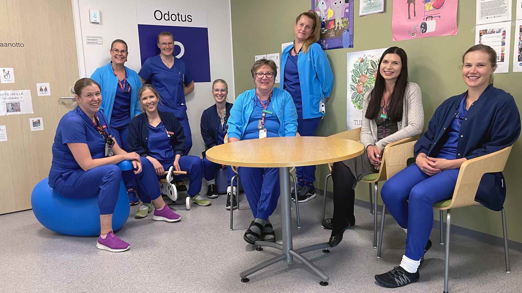 Yhdeksän terveydenhuollon työntekijää istuu ja seisoo odotustilan pöydän ympärillä. Taustalla näkyy odotustilan seinäopasteita ja julisteita.