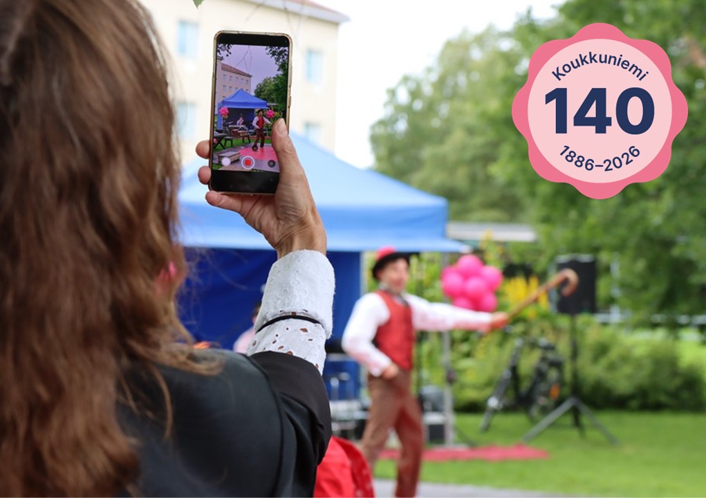 Henkilö ottaa kännykällään videota, ja taustalla näkyy sumeana Circus Simon. Yläkulmassa on Koukkuniemi 140 -leima.
