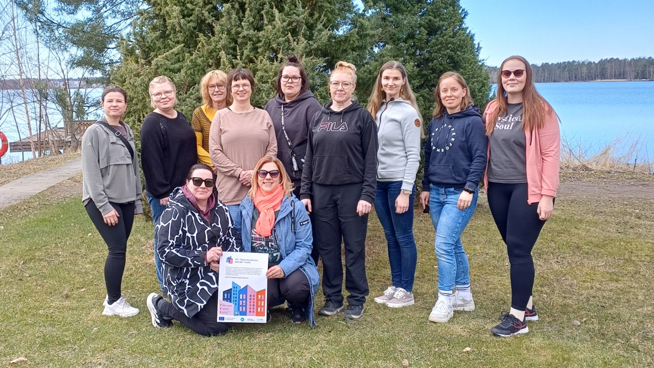 Työryhmän jäsenet seisovat järven rannalla nurmikolla ja poseeraavat kameraan. Etummaisilla on kädessään TIE-hankkeen esite.