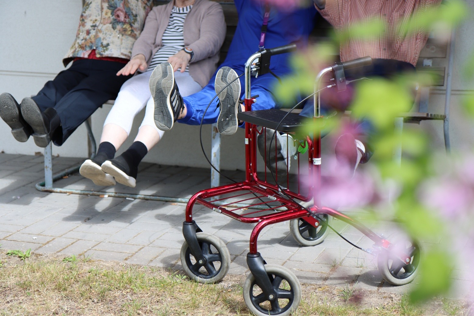 Ikääntyneet ja yksi hoitaja istuvat ulkona penkillä ja nostelevat jalkojaan ja käsiään. Edustalla näkyy myös rollaattori sekä sumeasti kukkapuuta.