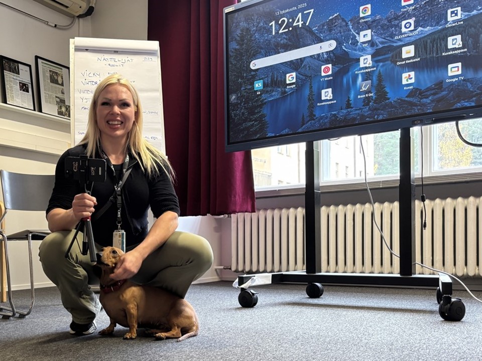 Ohjaaja Elina Oinonen on kyykyssä lattialla rapsuttamassa koiraa. Elinan oikealla puolella näkyy iso tietokoneen näyttö.