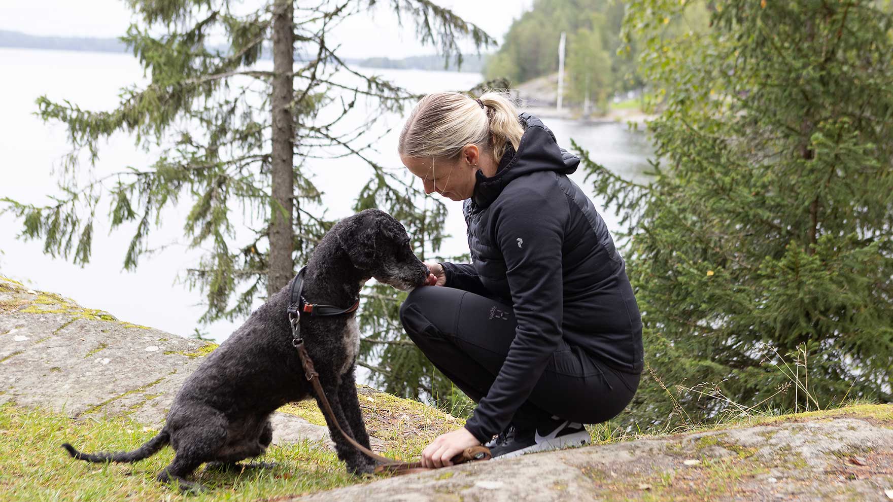 Nainen ja koira ovat vastatusten kyykyssä kalliolla, taustalla näkyy järvi.