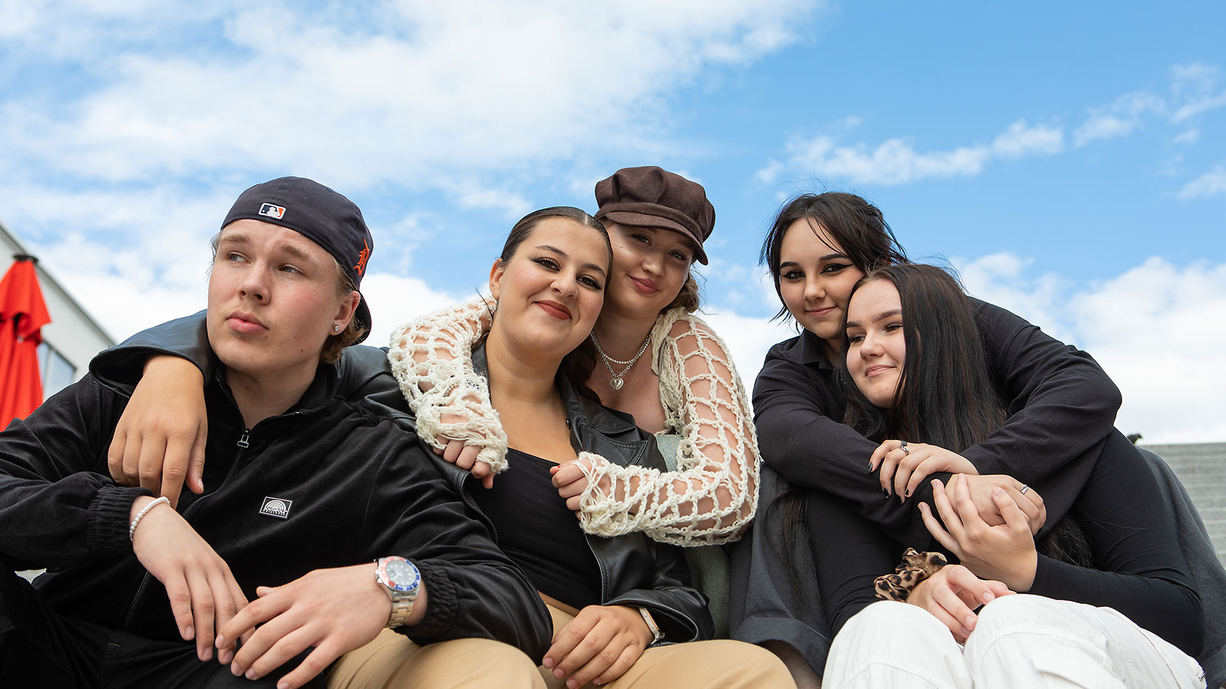 Five young people smiling and hugging each other, with a blue sky in the background.