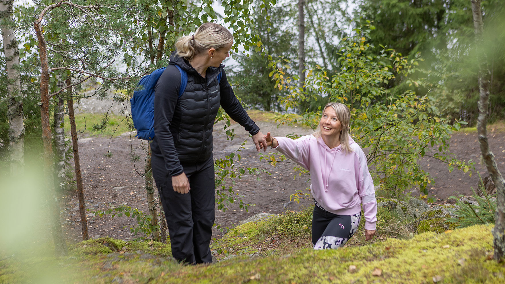 Kaksi naista metsässä ulkoilemassa, toinen nainen auttaa kaveriaan kädestä mäelle.