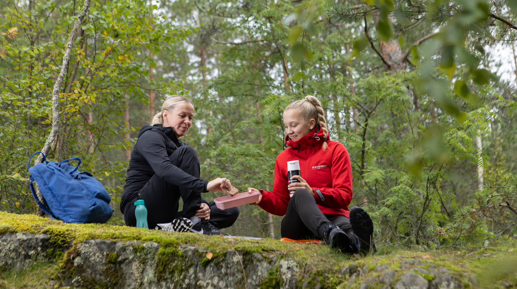 Kaksi henkilöä istuu sammaleisella kalliolla ja nauttii retkieväistä.