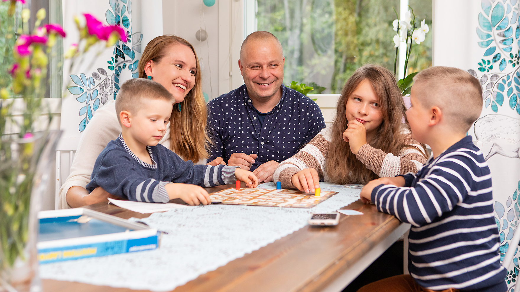 Kaksi aikuista ja kolme lasta pelaavat lautapeliä pöydän ääressä.