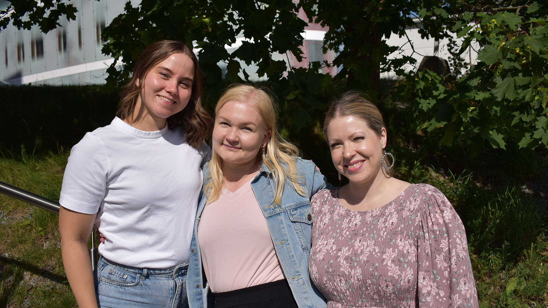 Kolme hymyilevää naista seisovat rinnakkain ulkona portaikon kaidetta vasten. Taustalla on kesäinen vaahtera.