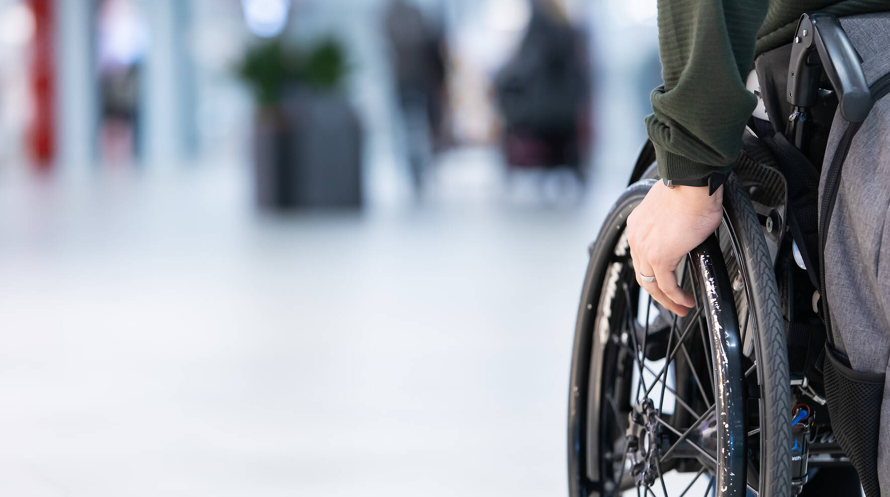 Kuvassa näkyy pyörätuolissa istuvan henkilön käsi ja pyörätuolin rengas. Taustalla on sumeana sairaalan käytävä.