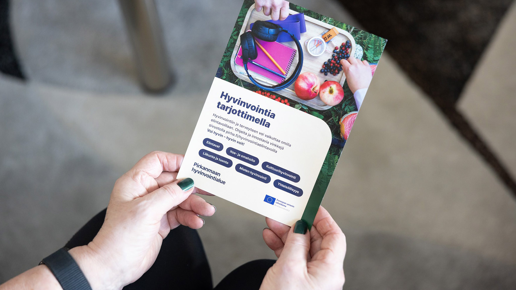 Henkilö pitää käsissään esitettä, jossa lukee Hyvinvointia tarjoittimella.