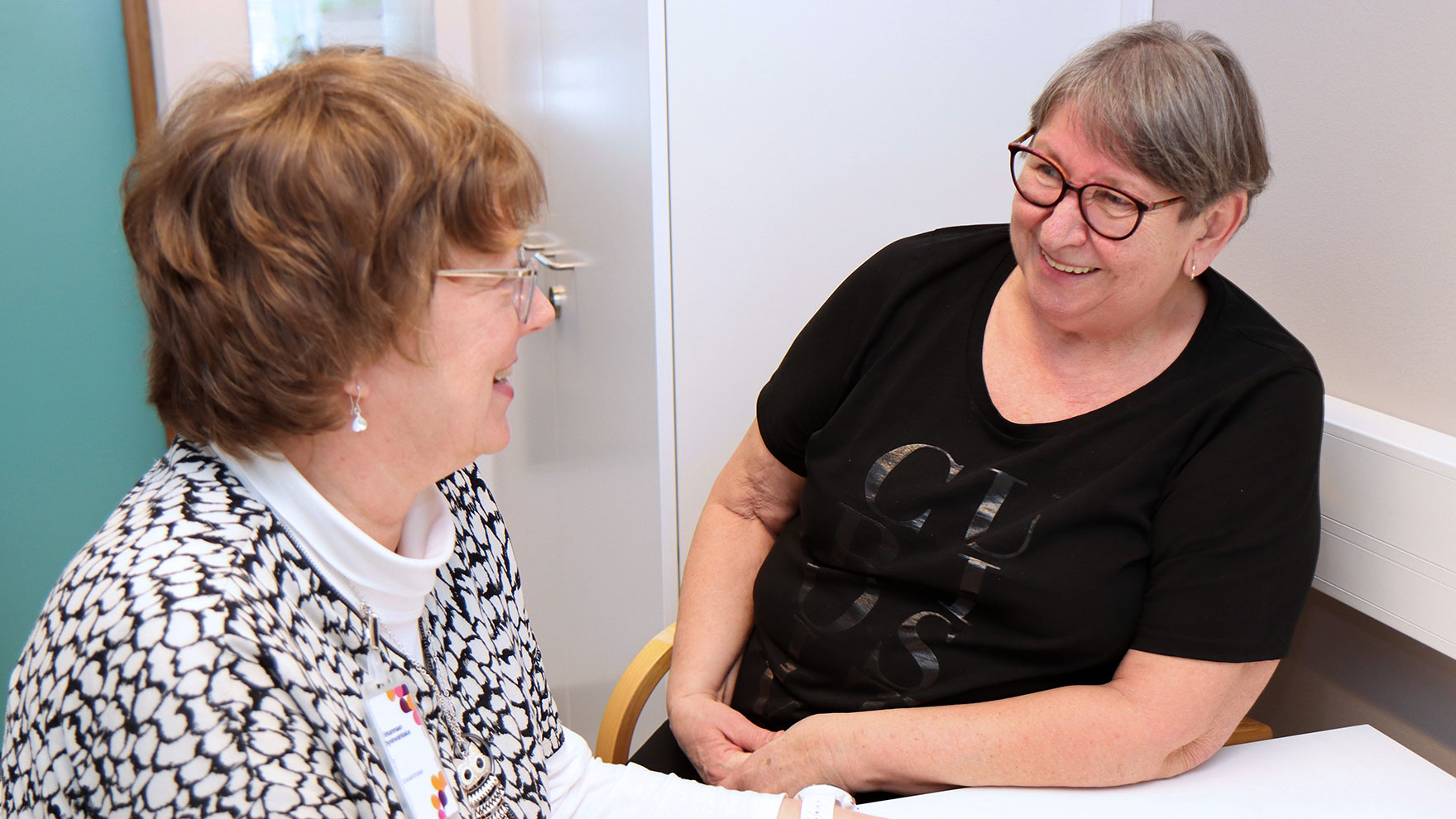 Kaksi naista keskustelee hymyillen pöydän ääressä. Toisella heistä on nimikortti, joka kertoo, että hän on Pirhassa töissä.