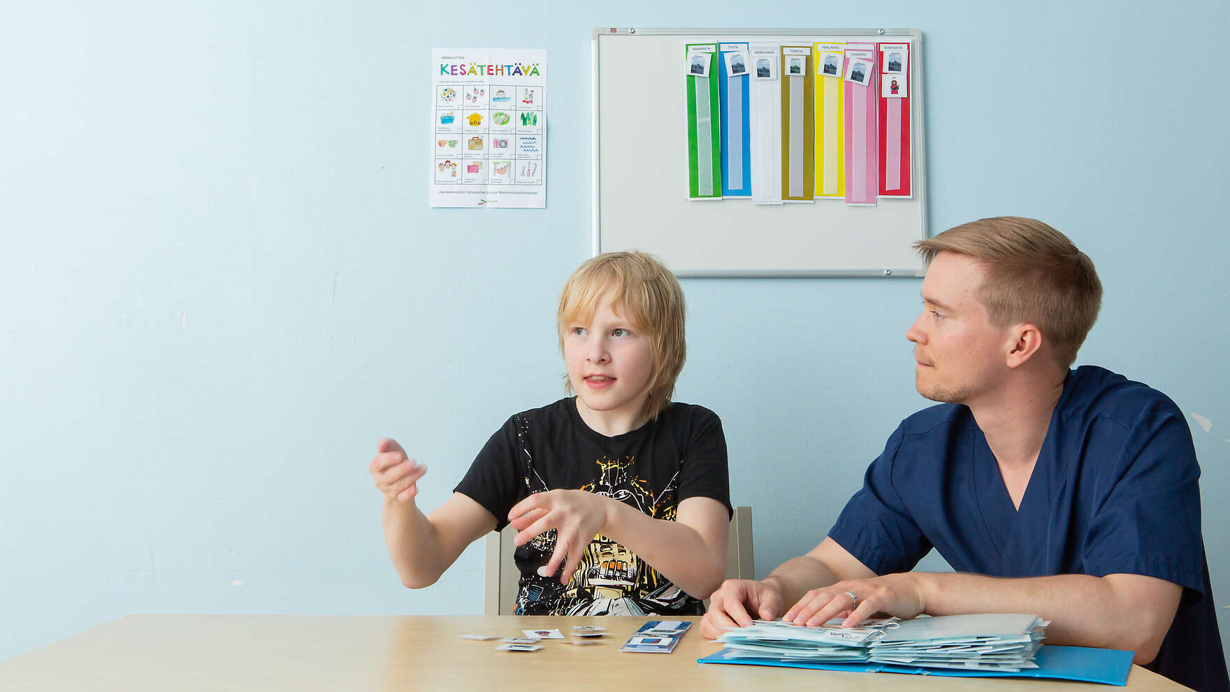 Lapsi ja hoitaja istuvat pöydän ääressä ja tekevät tehtäviä hoitokorteilla.