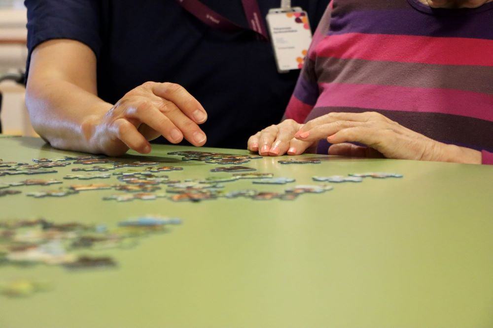 Pöydällä näkyy työntekijän ja ikääntyneen kädet sekä palapelin paloja.