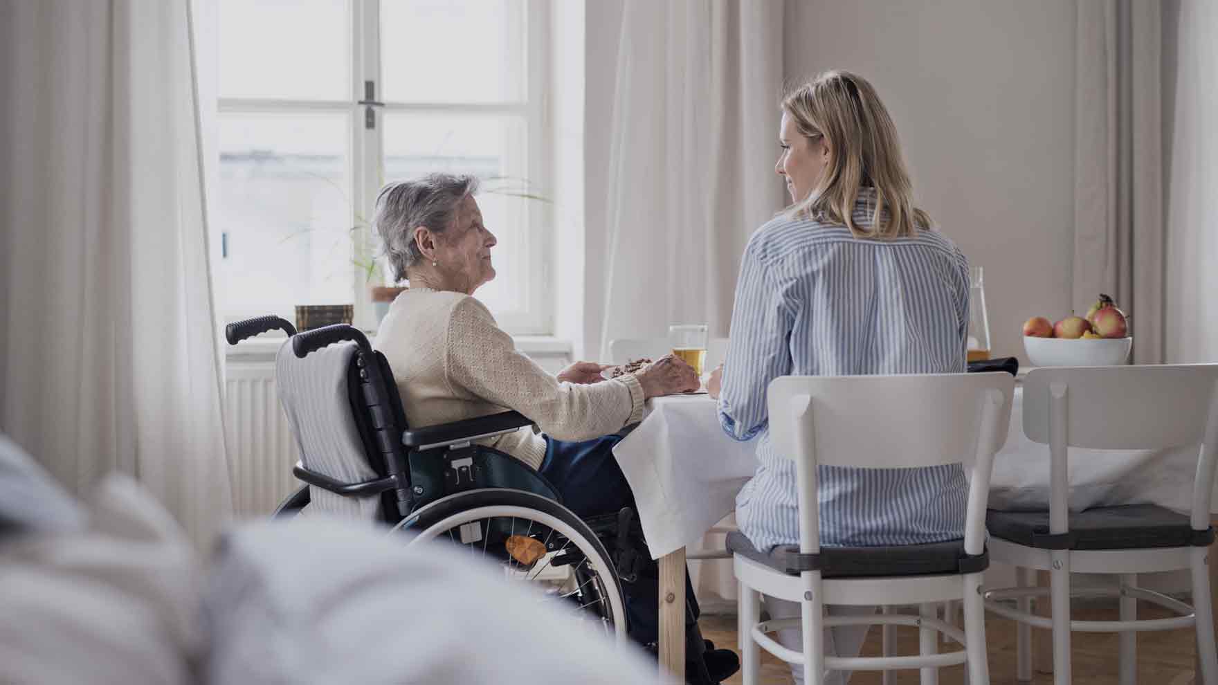 Ikääntynye nainen istuu pyörätuolissa pöydän ääressä. Hänen vieressään selin kameraan istuu nuori vaaleatukkainen nainen. Pöydässä on ateria.