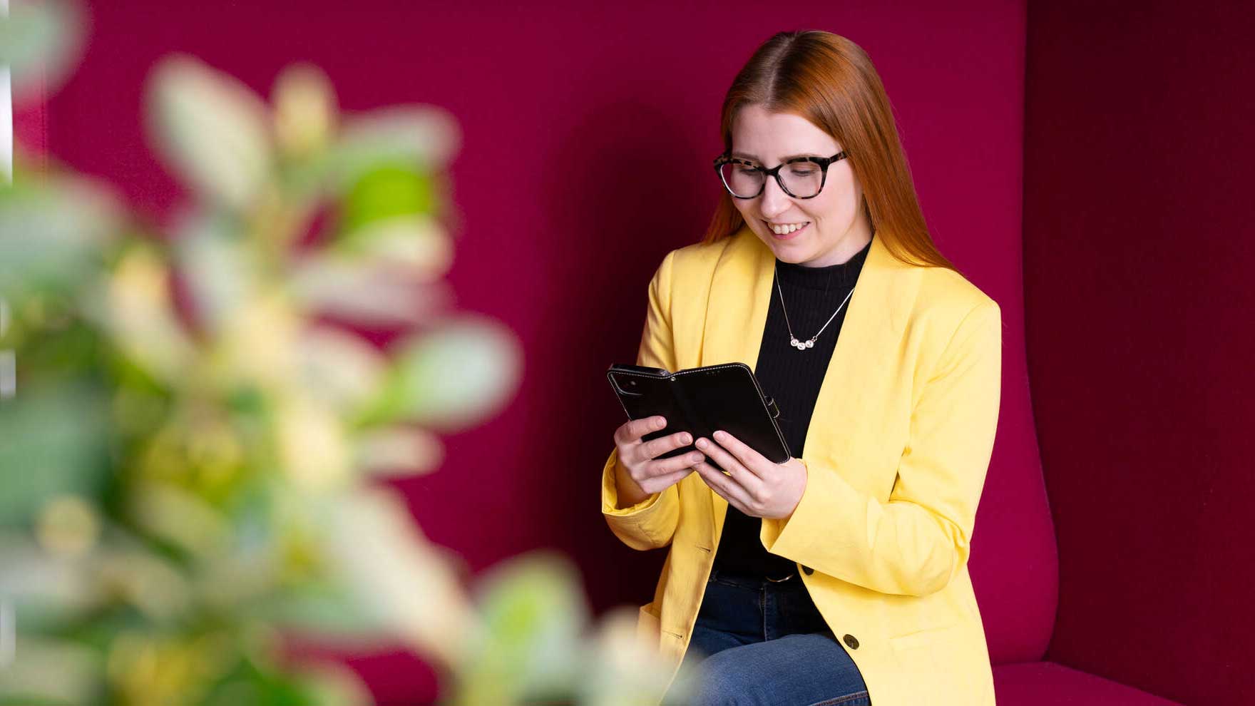 A woman in a yellow jacket sits on a red sofa and looks at her mobile phone.