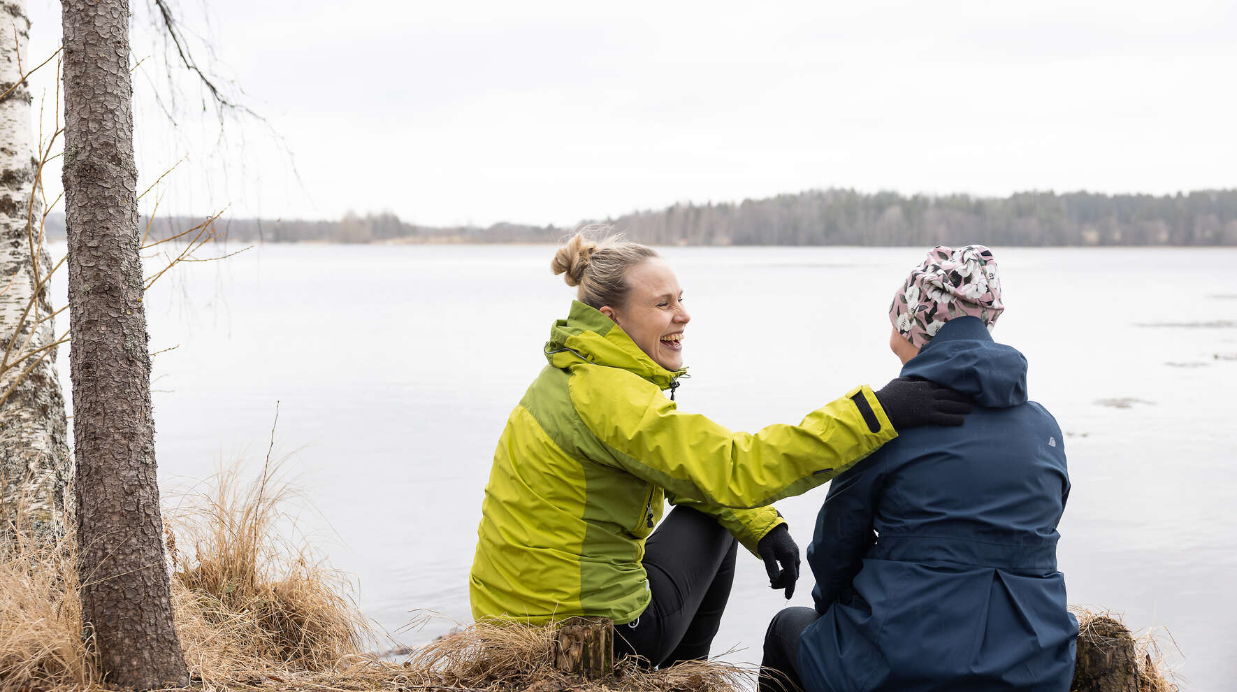 Kaksi naista istuu rantapenkalla, toinen pitää toista olkapäästä ja nauraa.