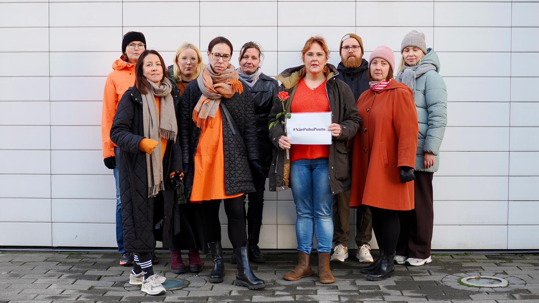 Yhdeksän henkilön työyhteisö, joista yksi pitää kädessään #NäePuhuPuutu -kylttiä.