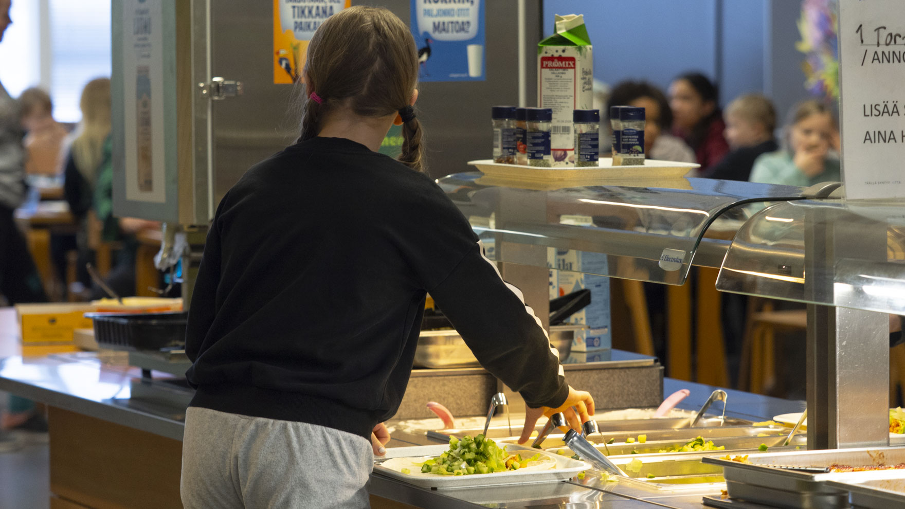 Kuva kouluruokalinjastosta, jossa oppilas selin kameraan ottamassa ruokaa.