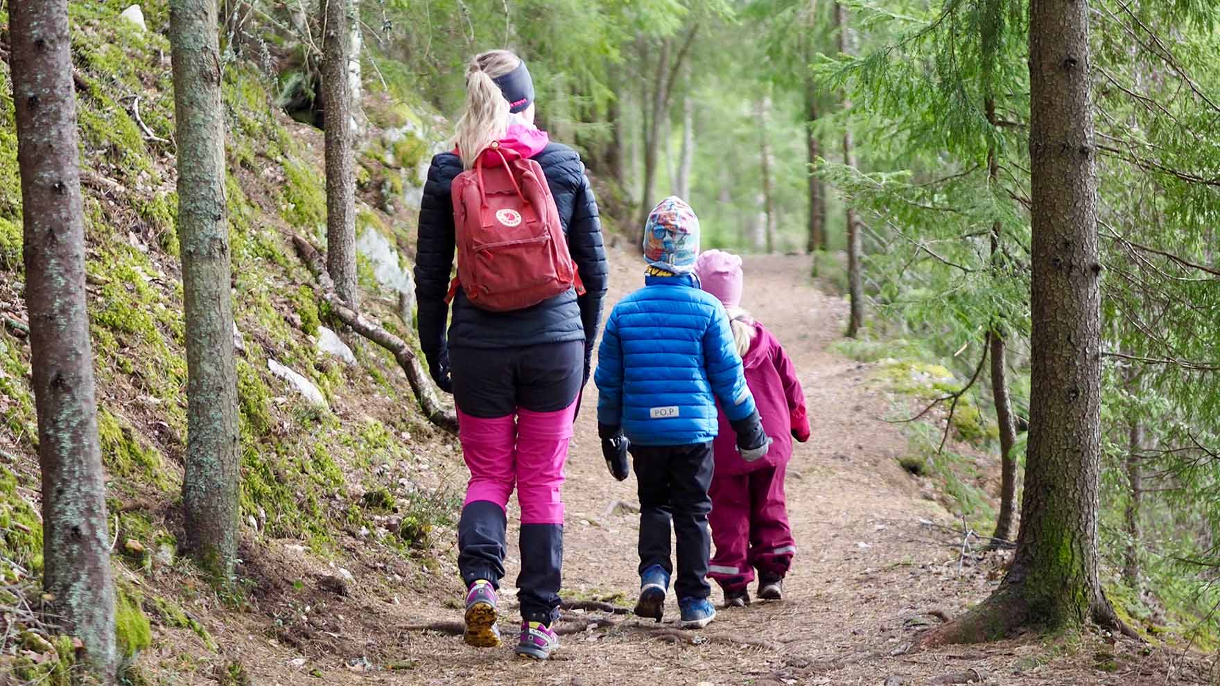 Aikuinen nainen ja kaksi alle kouluikäistä lasta kävelevät metsäpolulla pois päin kamerasta.