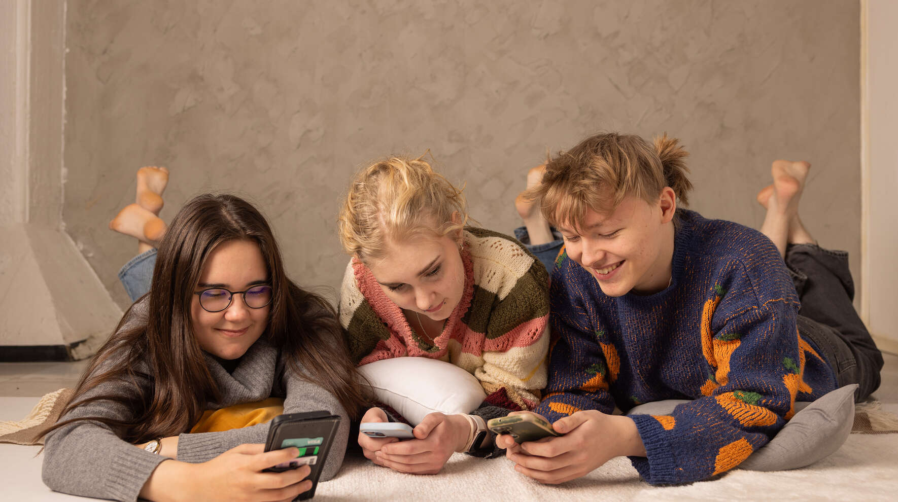 Kolme nuorta makaa rinnakkain vatsallaan ja katsoo puhelimiaan.
