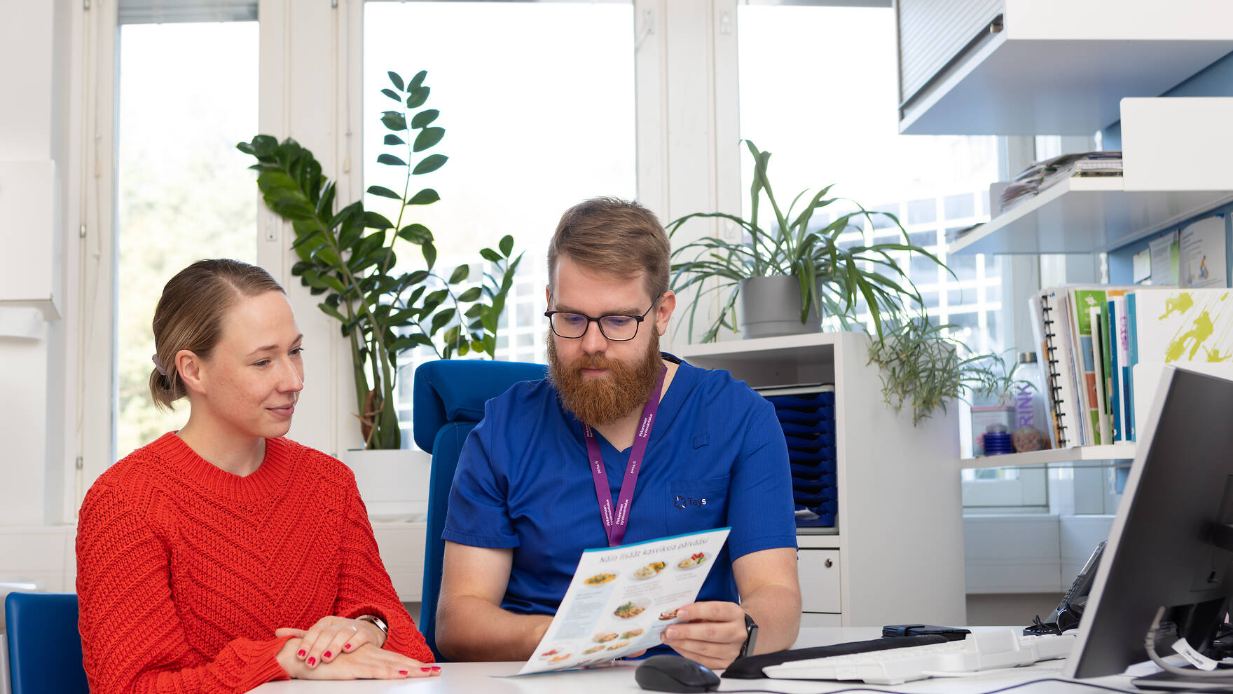 Terveydenhuollon ammattilainen ohjeistaa asiakasta vastaanottotilan pöydän äärellä.