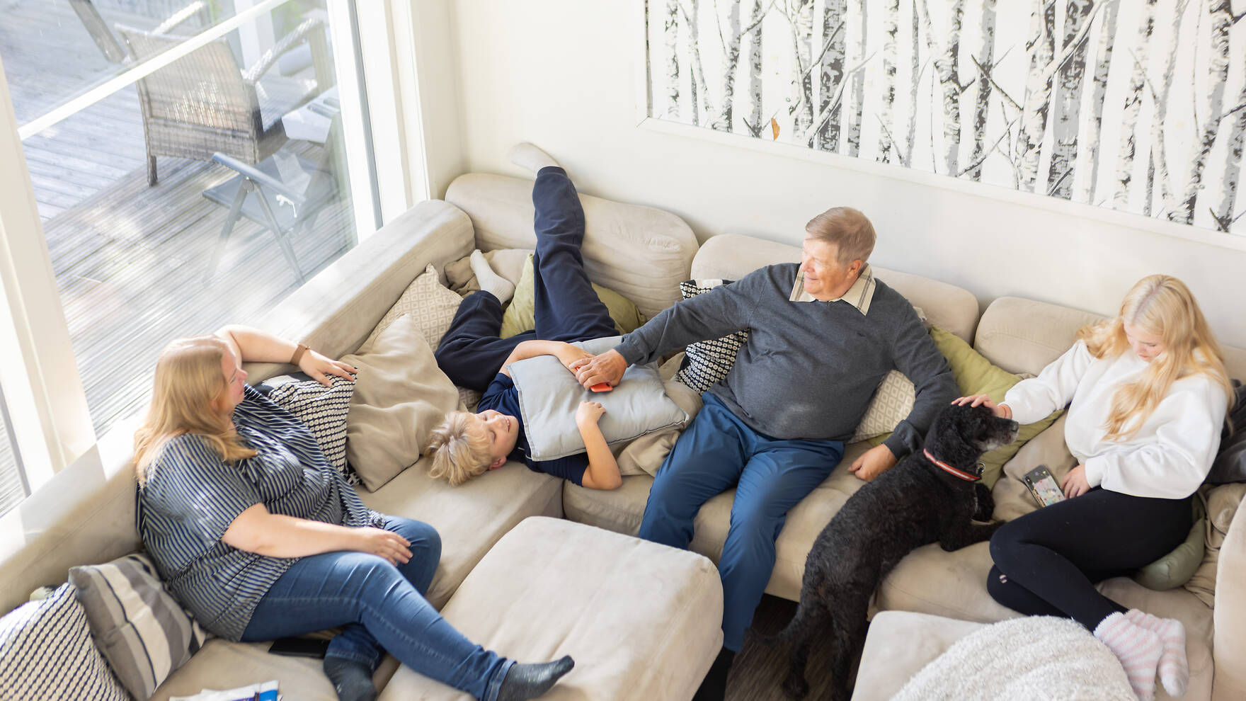 Yläviistosta otettu kuva, jossa vaalealla sohvalla istuu ikääntynyt mies ympärillään nuori poika ja poika sekä keski-ikäinen nainen.