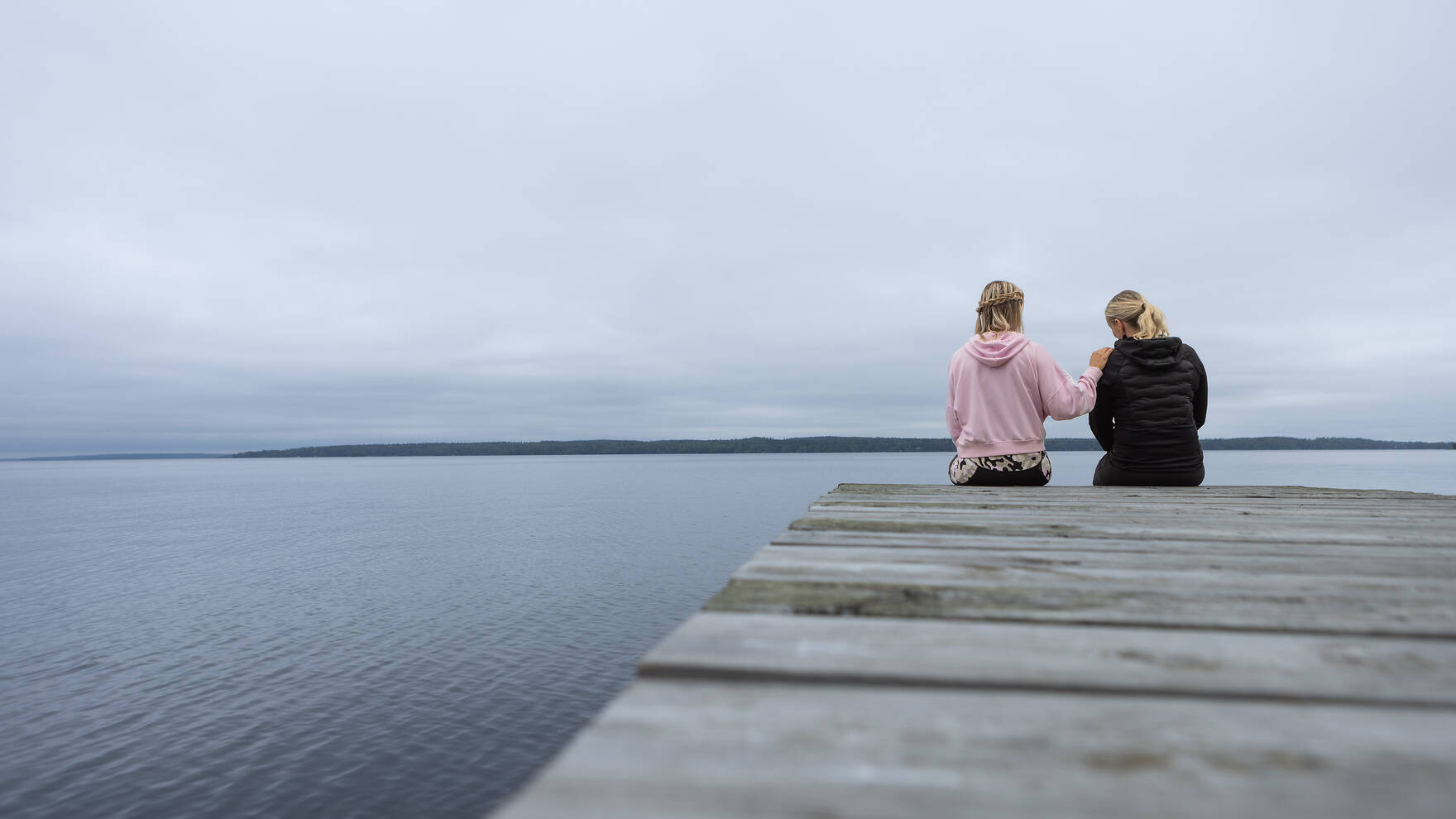 Kaksi naista istumassa laiturin päässä ja toisen käsi lohduttavasti toisen olalla.