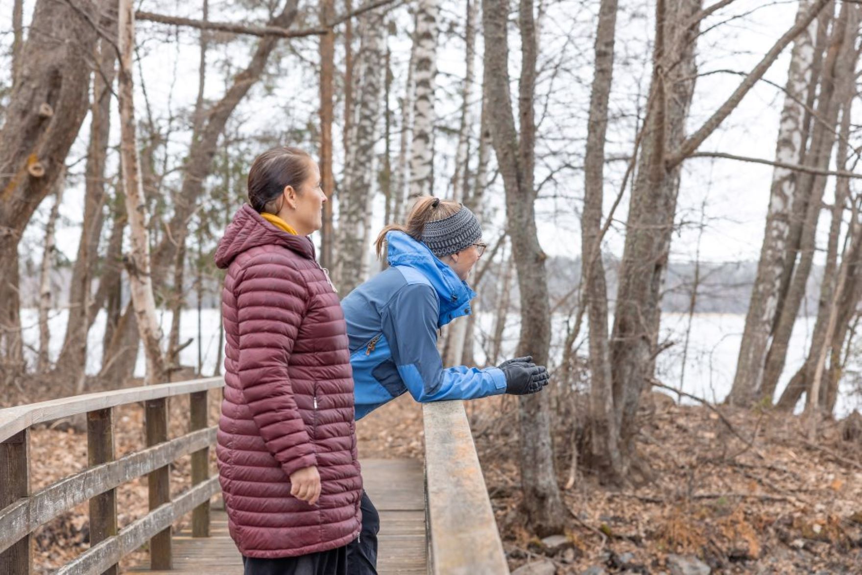 Kaksi henkilöä nojailee puisella sillankaiteella katsellen puiden takana näkyvää järveä.