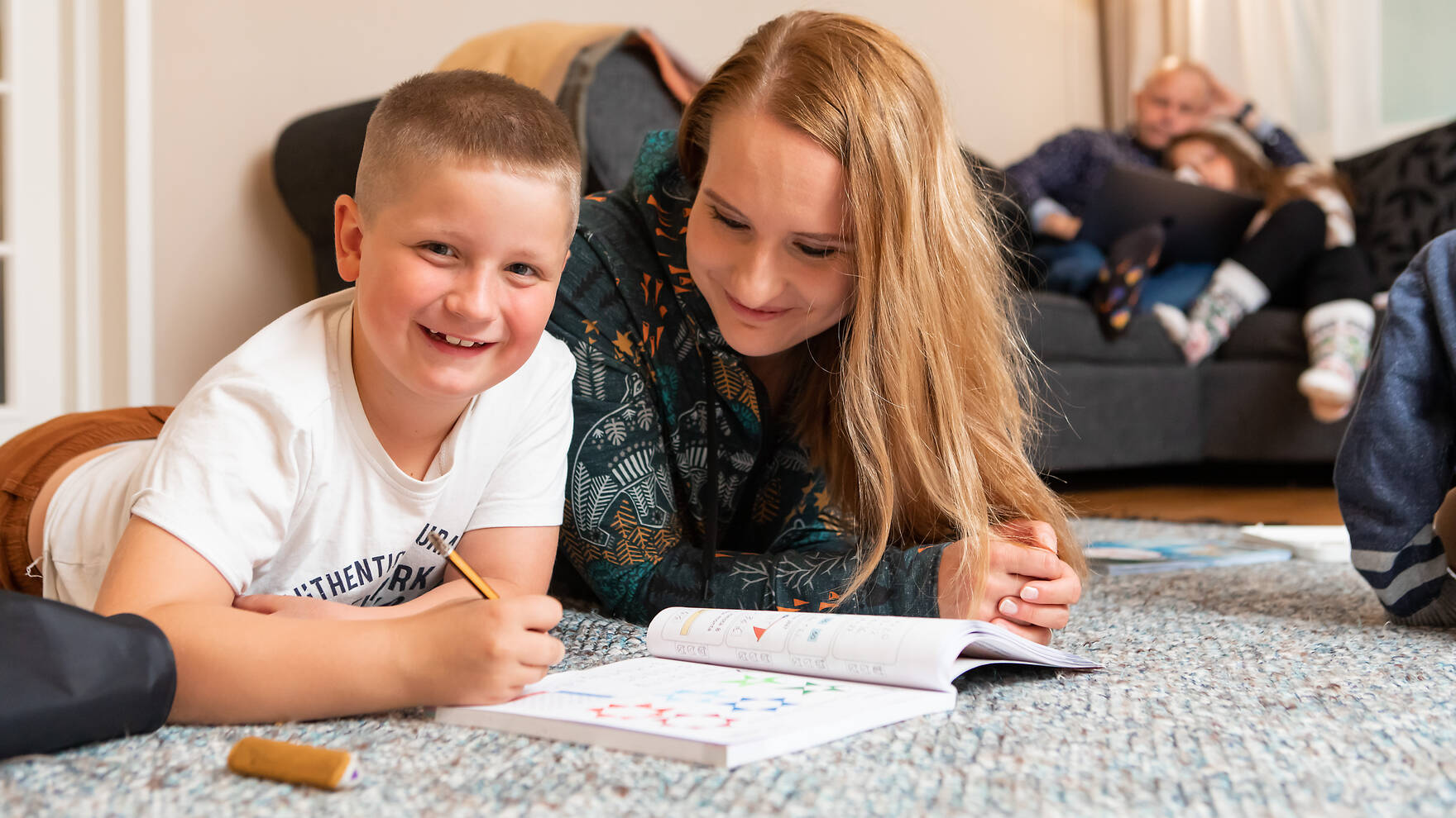 Äiti tukee poikaa läksyjen tekemisessä. Taustalla isä ja tyttö istuvat sohvalla kannettavan tietokoneen äärellä..