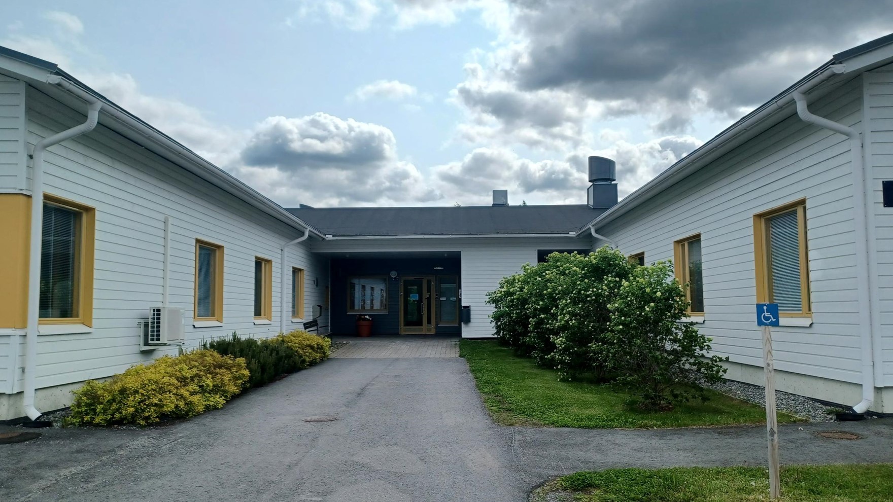 Vaaleasta laudasta tehty matala rakennus ja sen sisäänkäynti, jossa istutuksia