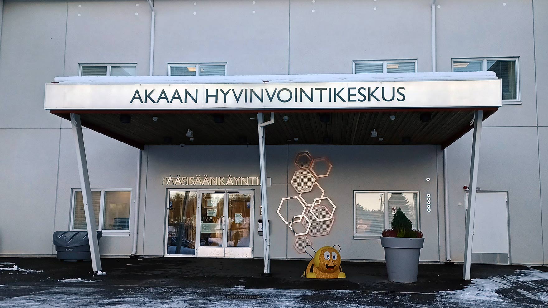 Valkoisen talon edessä on katos, jossa lukee "Akaan hyvinvointikeskus". Katoksen alla on hyvinvointikeskuksen pääsisäänkäynti.