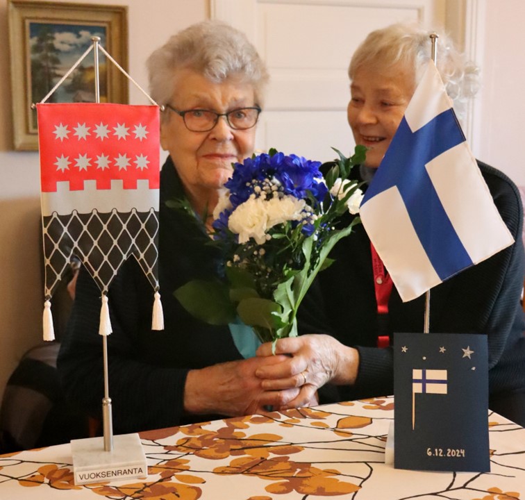 Veteraani Maija ja vapaaehtoinen Riitta katsovat kameraa, Maijalla on kädessään veteraaneille toimitetut kukat.