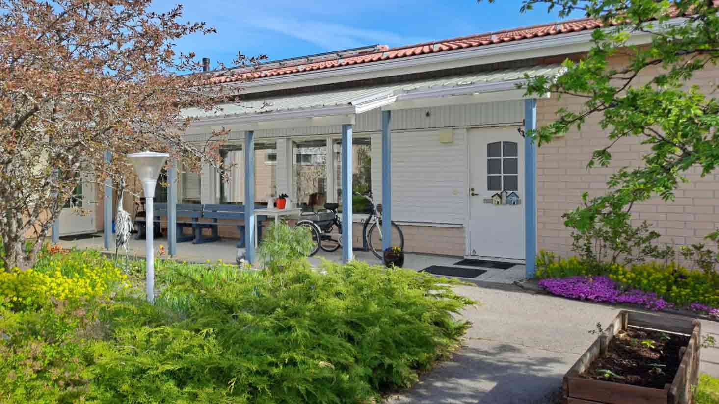 Vaaleasävyinen rakennus, jonka edessä on istutuksia ja kolmipyöräinen polkupyörä.