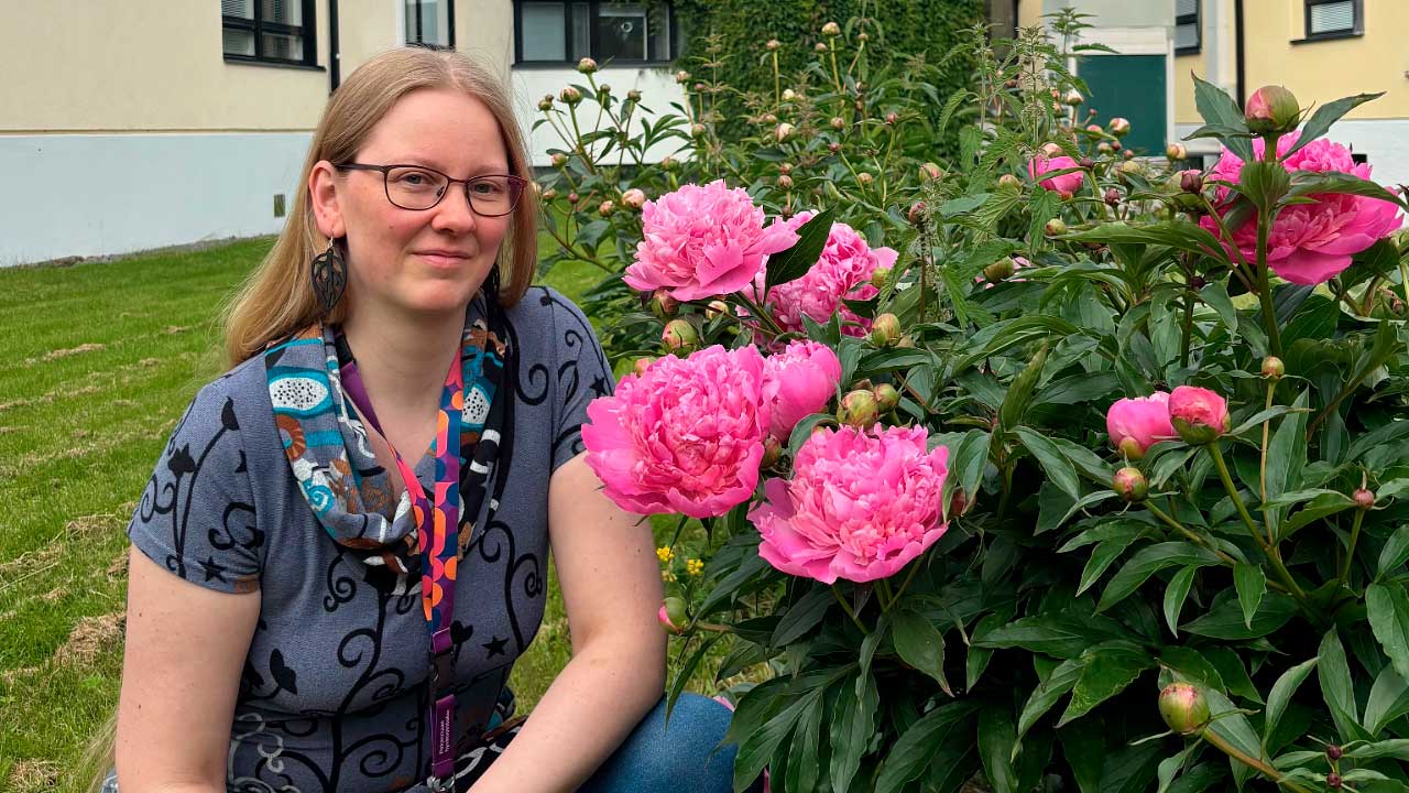 Jaana istuu pionipuskan vieressä kesäisissä maisemissa. 