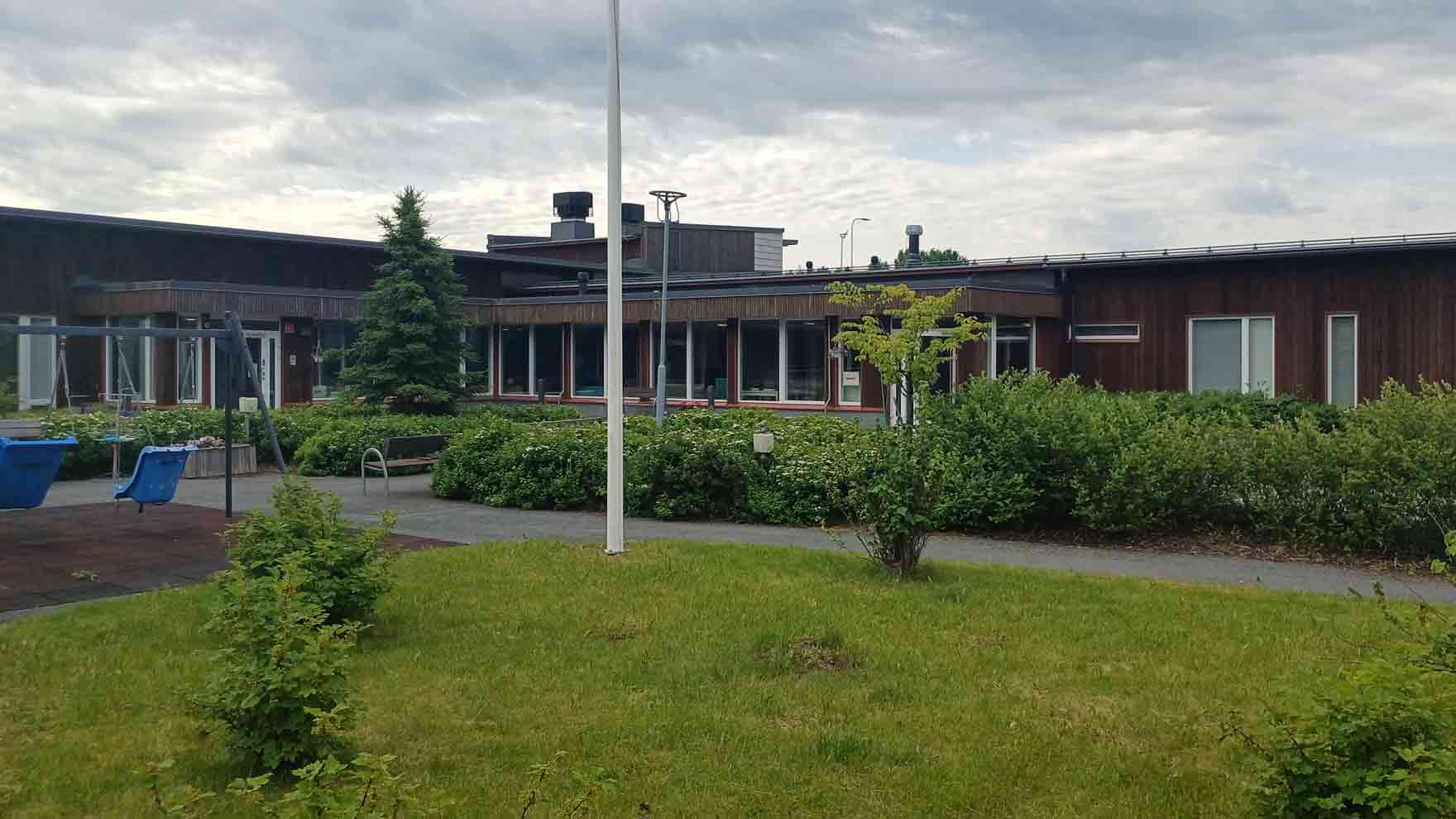 Punertavan sävyinen rakennus, jonka edessä paljon istutuksia ja keinu