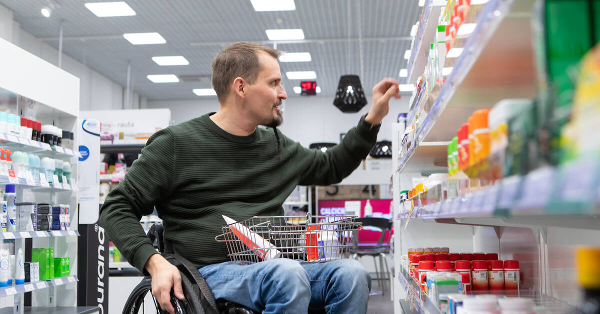 Pyörätuolilla kulkeva asioi apteekissa ja on ottamassa hyllyltä tuotteita.