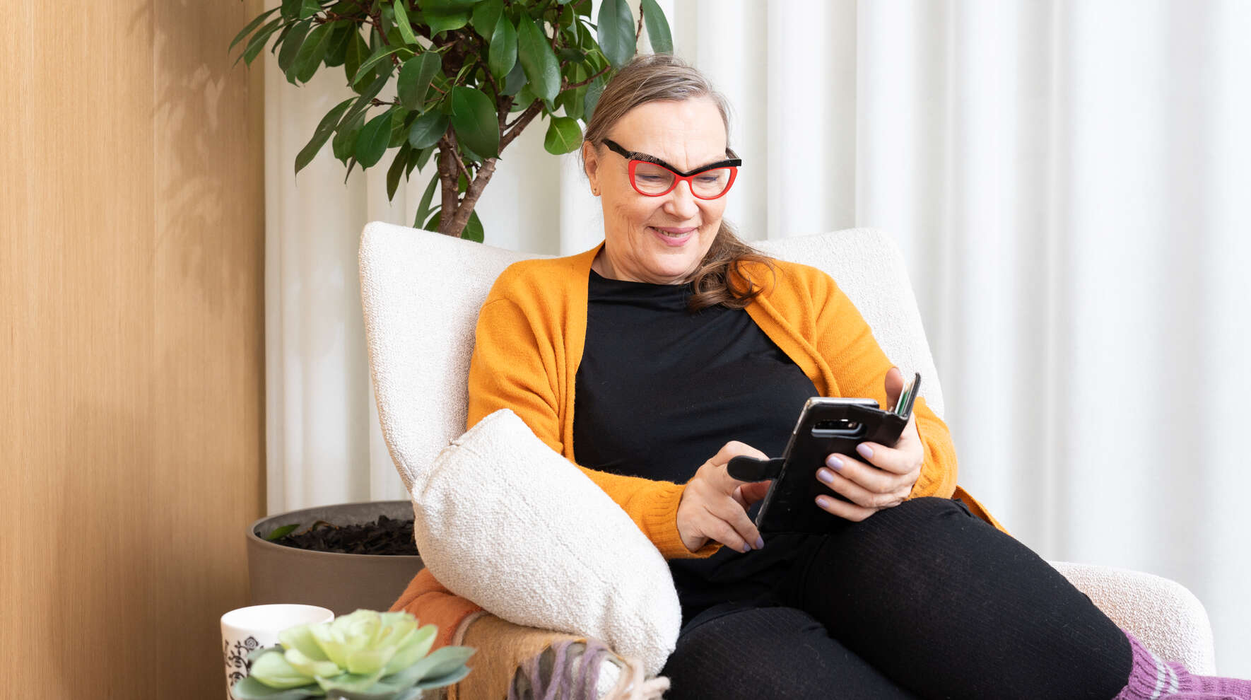 Hymyilevä nainen istuu nojatuolissa ja näppäilee puhelinta.