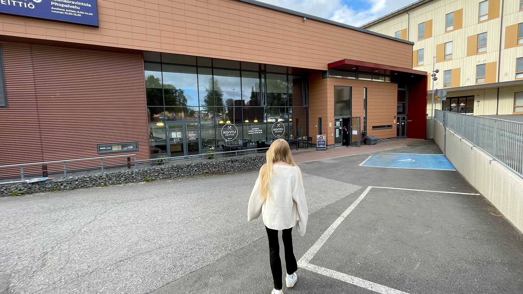 Nuori kävelee asfalttipihan poikki kohti vaaleanruskeaa rakennusta.