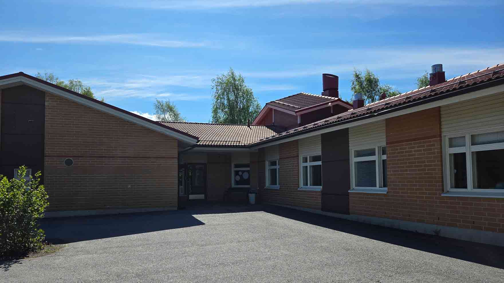 Pääosin punatillinen matala rakennus, jonka edustalla tasainen piha