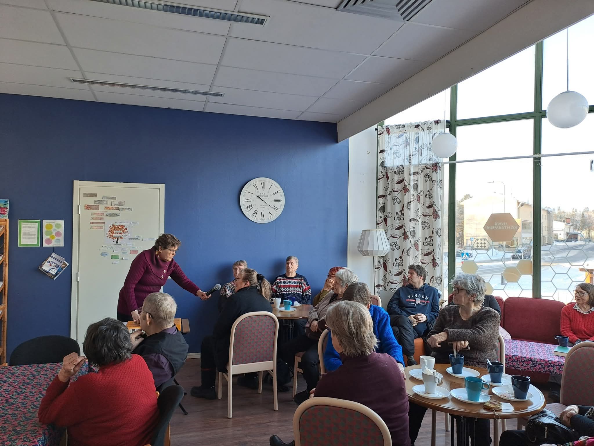 Ihmiset istuvat penkeillä ja seuraavat tapahtumaa. Ihminen ojentaa mikrofonia yleisöä kohti.