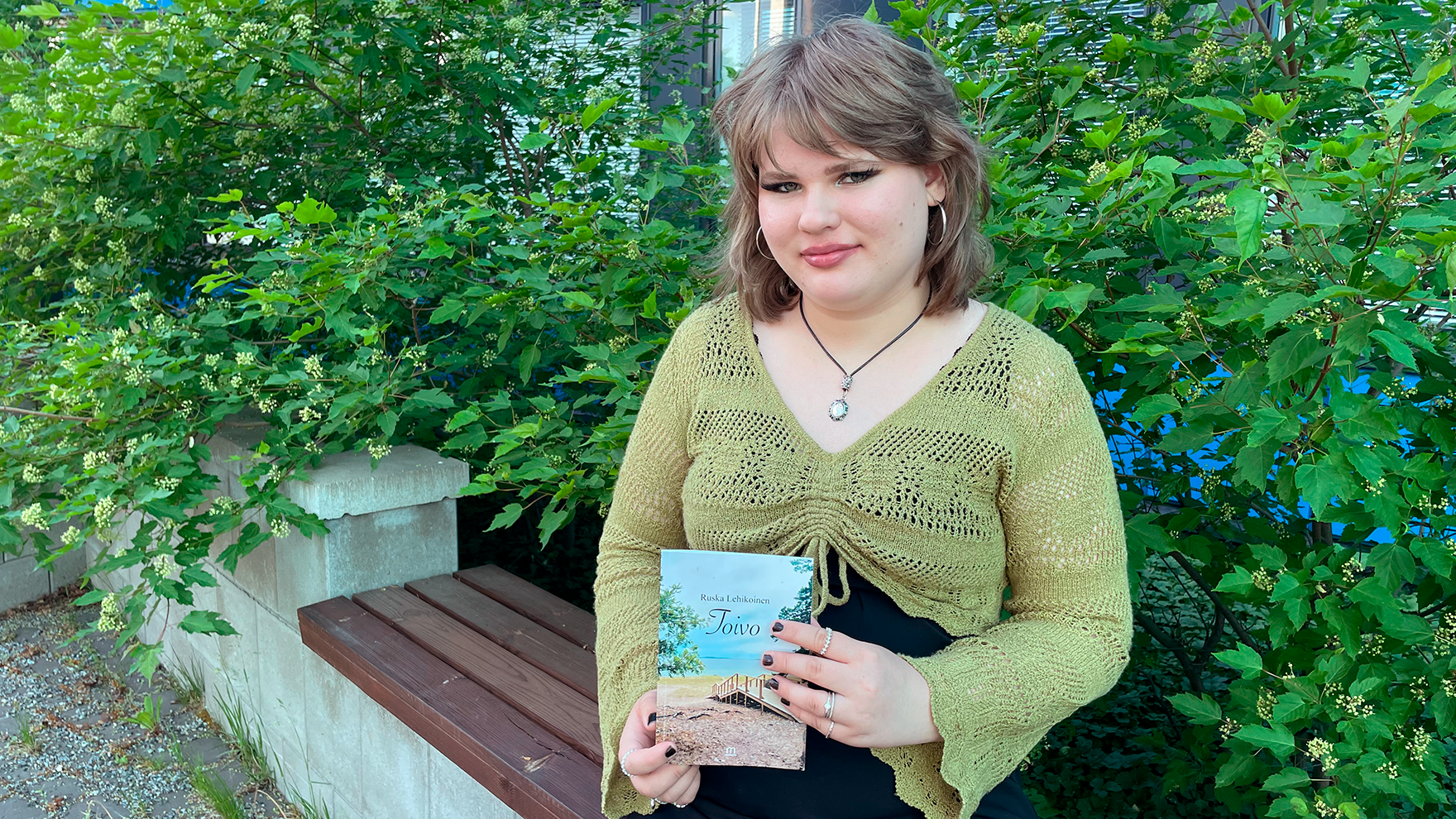 Kuvassa Ruska Lehikoinen istuu penkillä ja pitelee käsissään Toivo-kirjaa, jonka hän on kirjoittanut. Taustalla on vihreitä pensaita.