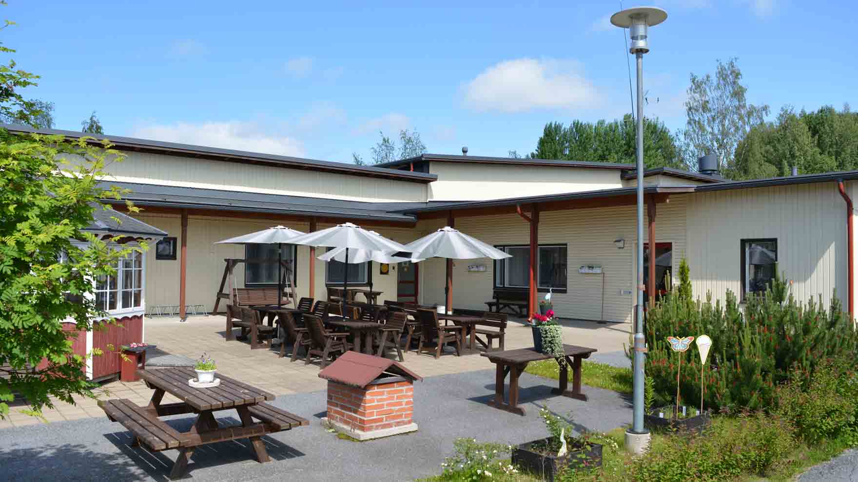 Vaalea matala rakennus, jonka sisäpihalla pihatuoleja ja aurinkovarjoja