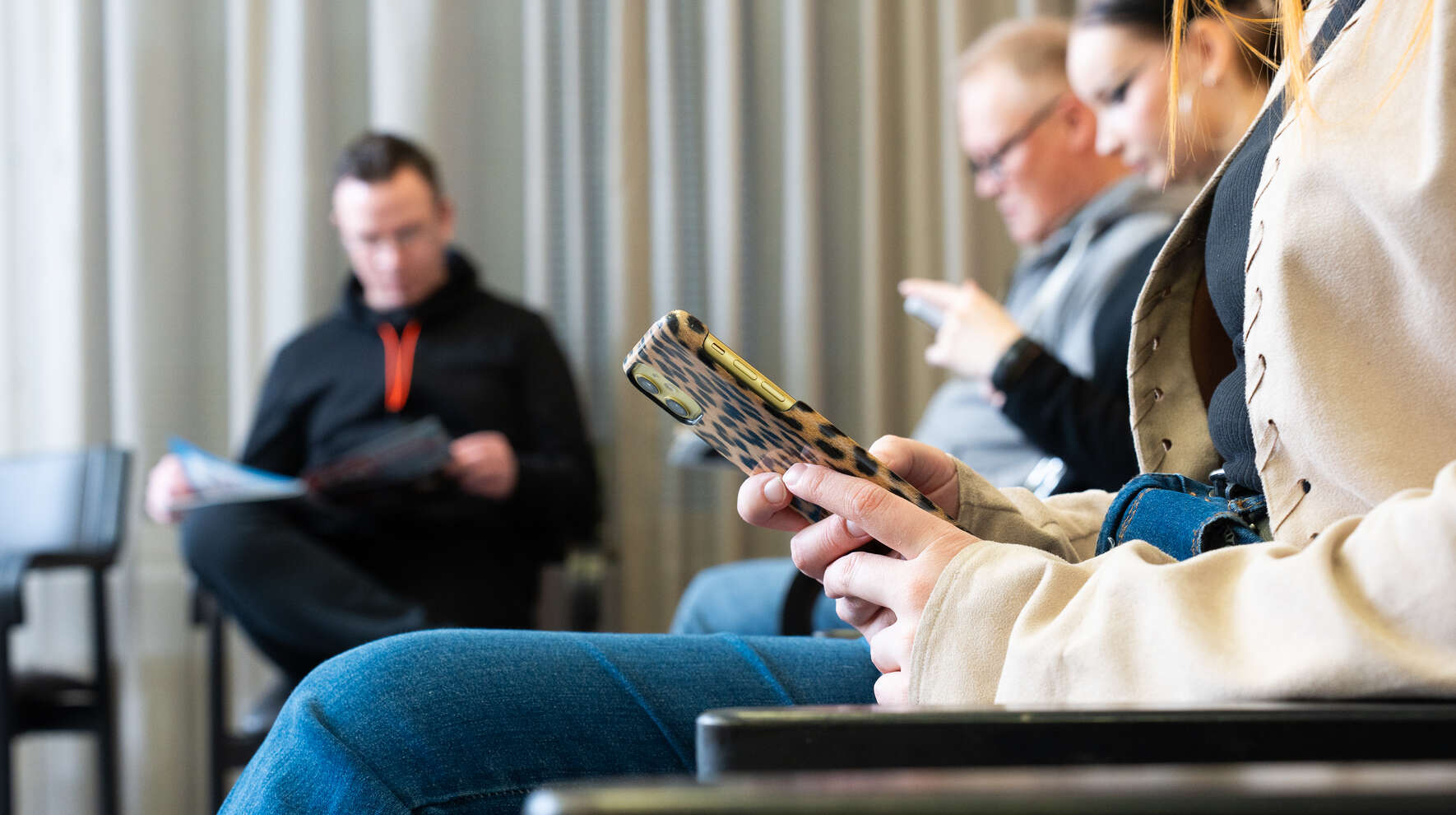 Neljä henkilöä istuu odotushuoneessa ja lukee lehtiä tai selaa puhelinta.