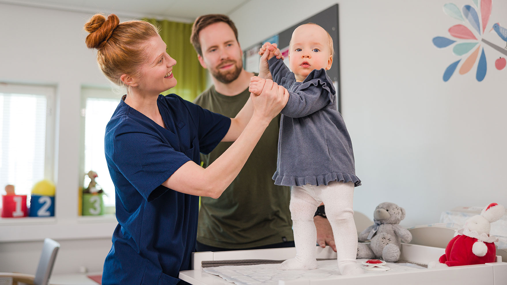 Isä ja pieni lapsi neuvolakäynnillä. Lapsi seisoo hoitopöydällä ja hoitaja kannattelee häntä käsistä.