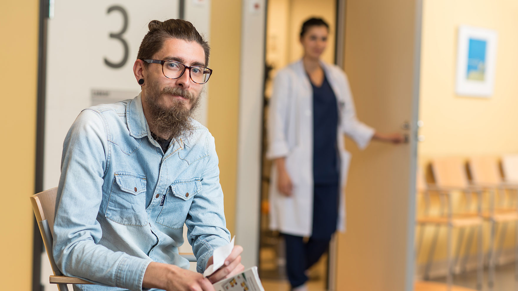 A man is sitting in the lobby of a health centre, looking towards the camera, with a health professional standing in the background at the door of the reception room.