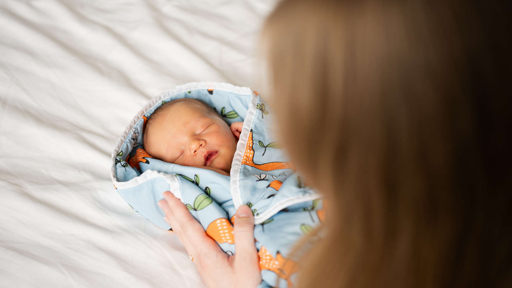 Vauva on nukkuu selällään käärittynä värikkääseen peittoon ja aikuisen kädet ovat peiton päällä.