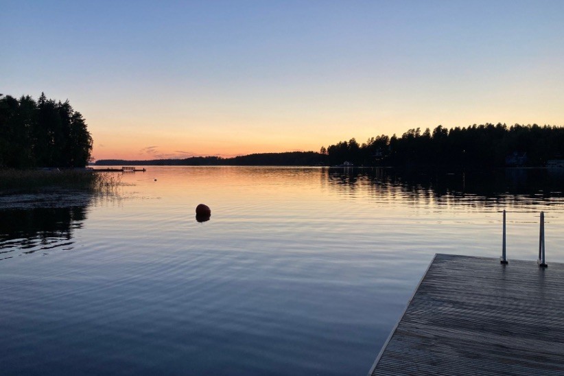 Loppukesän järvimaisema jossa näkyy laituri, tyyni järvi ja juuri laskenut aurinko horisontissa metsän takana.